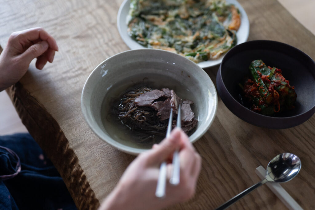 平壌冷麺、チヂミ、オイキムチの夏膳を前に箸を取る